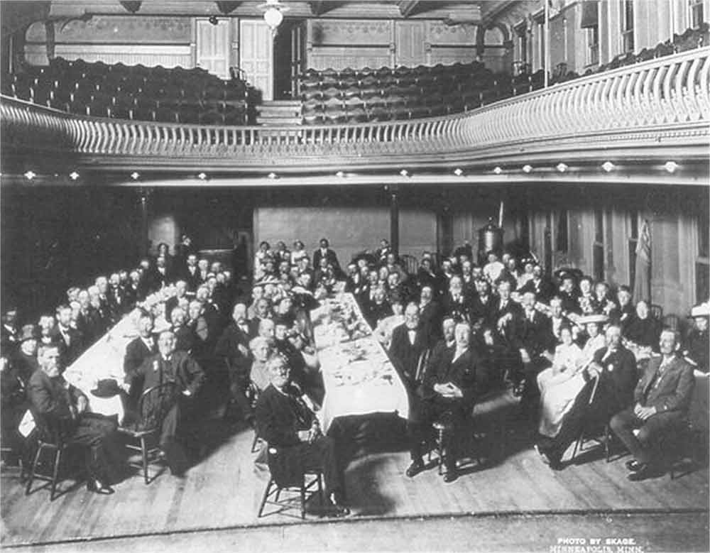 Two story hall with empty seats in balcony. Floor contains three long, parallel tables with people (mostly men) on both sides of each table dressed formally in 1910 attire.