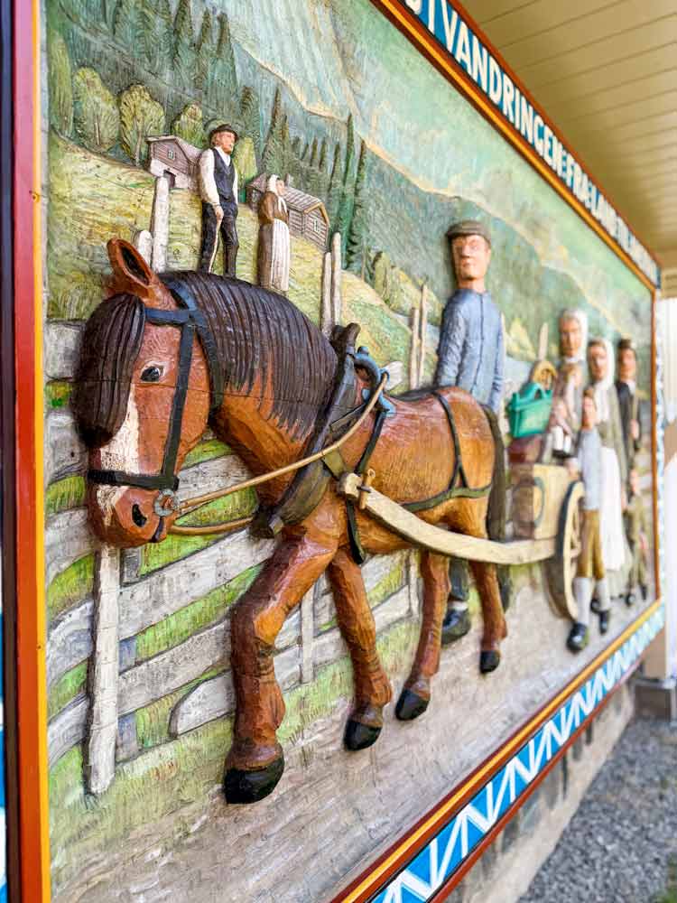Painted, wood carving on a big surface (maybe 14' wide by 6 feet high) of Norwegian emigrants in Norway leaving with a cart and horse walking by friends who are staying with a border around it with words translated from Norwegian as "Remembrance of the Emigration From Land To America". Picture is close-up, taken from an angle.