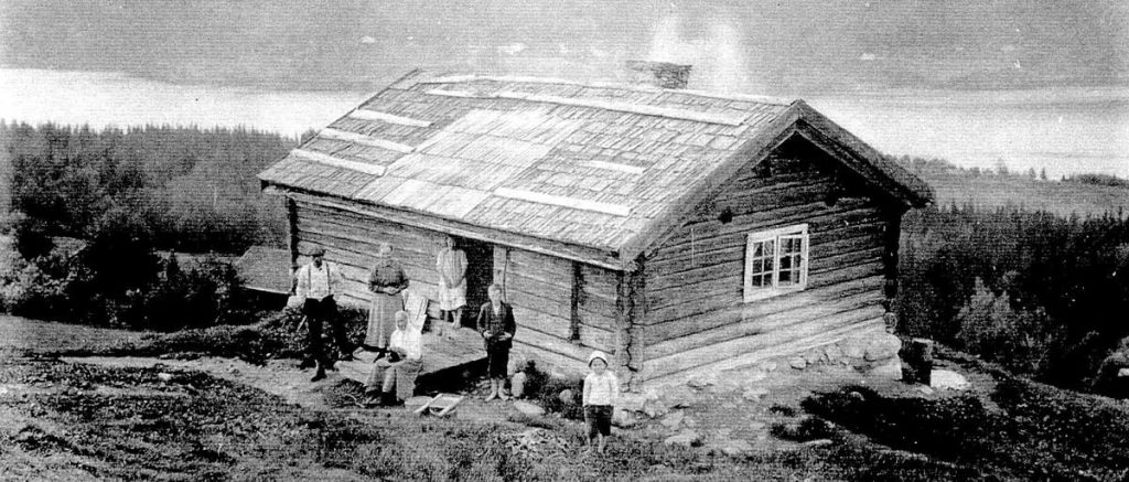 Vintage picture of a husmannsplass (cottage) sitting high on a hill. It has people posing in front and the Randsfjord below in the background.